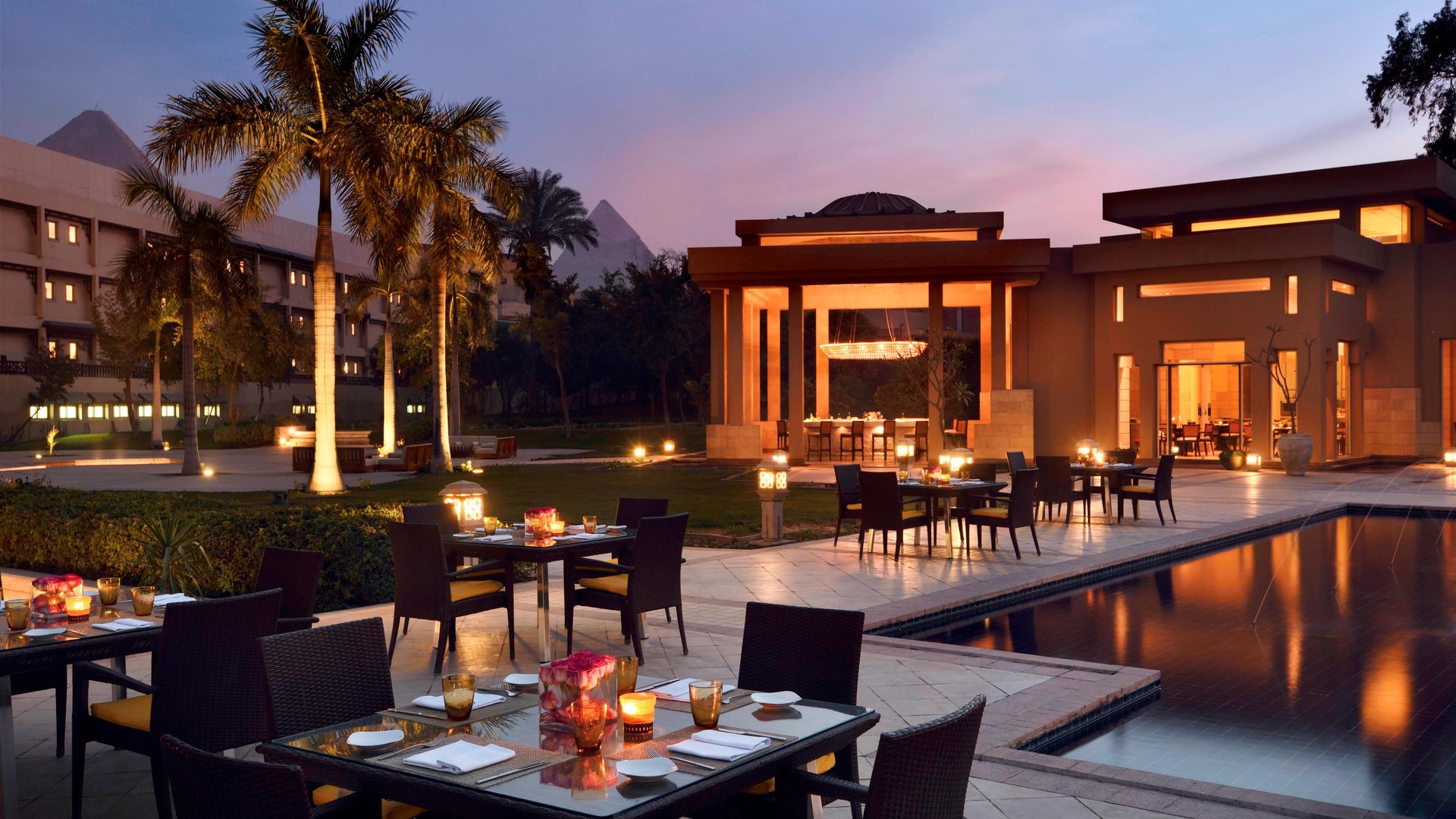 Tables set by the pool at Mena House at dusk, with the Pyramids behind the palms